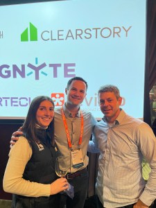 A Remy team member with two attendees at a partner event, in front of a screen of construction tech partner logos