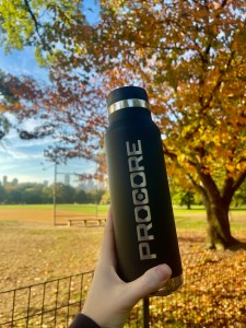 A Procore water bottle held up in a park with autumn trees